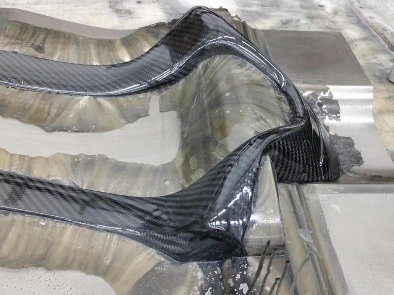 Image shows the head and neck safety device in its tool in a production environment. The composite bladder molded structure has not been removed from the tool and there is resin flashing on the tool along the edges of the hardware where the tool halves met. The surface of the hardware is a high gloss carbon fiber weave.